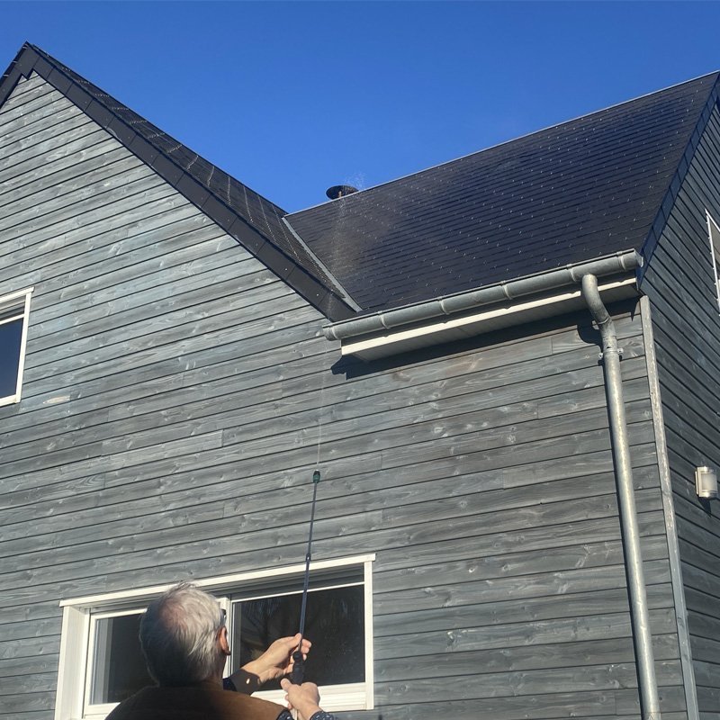 Homme utilisant un pulvérisateur électrique Ecojet sur façade de maison à toit gris, fenêtre au premier plan et tuyaux visibles.